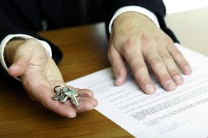 one hand of businessman gives  house keys to the hand of another businessman. Signed contract and pen visible in background.